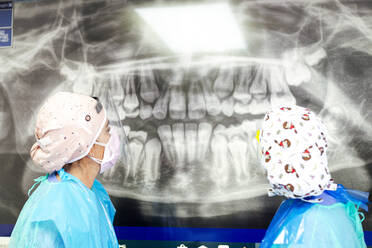 Zahnarzt und Assistentin in Arbeitsschutzkleidung bei der Untersuchung von Röntgenbildern in einer Zahnarztpraxis - JCMF01267