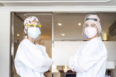 Confident doctor and assistant in protective workwear standing with arms crossed at office - JCMF01283