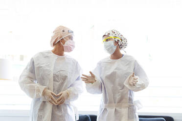 Doctor and assistant in protective suit talking while standing against window - JCMF01287