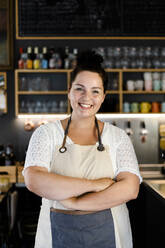 Smiling female owner with arms crossed standing in coffee shop - GIOF08760