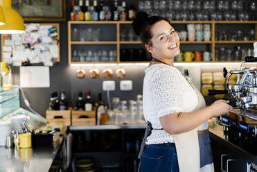 Lächelnde weibliche Barista, die bei der Arbeit in einem Café an der Kaffeemaschine steht - GIOF08762