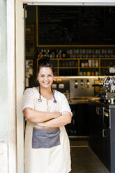 Smiling female owner with arms crossed standing at entrance of cafe - GIOF08766
