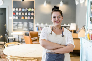 Lächelnde Besitzerin mit verschränkten Armen, die an den Möbeln eines Cafés steht - GIOF08768