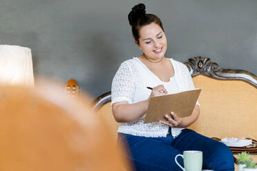 Lächelnde junge Frau schreibt in einem Buch und sitzt auf einem Stuhl in einem Café - GIOF08770