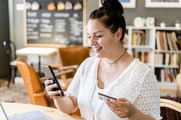 Close-up of smiling young woman holding credit card using smart phone in coffee shop - GIOF08776