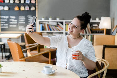 Smiling woman holding drink taking selfie with smart phone at table in cafe - GIOF08780