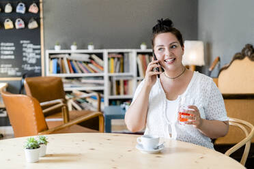 Üppige Frau, die ein Getränk hält und über ein Smartphone spricht, während sie in einem Café sitzt - GIOF08781