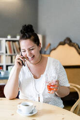 Fröhliche junge Frau, die ein Getränk in der Hand hält und über ein Smartphone spricht, während sie in einem Café sitzt - GIOF08782