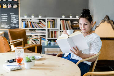 Junge Frau, die eine Kaffeetasse hält und ein Buch liest, während sie am Tisch in einem Café sitzt - GIOF08786