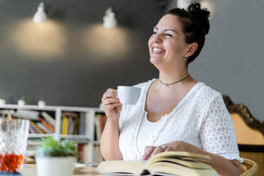 Fröhliche junge Frau schaut weg, während sie mit Kaffee und Buch in einem Café sitzt - GIOF08788