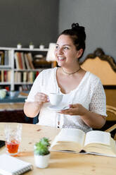 Glückliche junge Frau mit Buch auf dem Tisch, die eine Kaffeetasse hält, während sie im Restaurant sitzt - GIOF08789