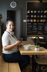 Fröhliche junge Frau, die ihr Smartphone benutzt, während sie am Tisch in einem Café sitzt - GIOF08790