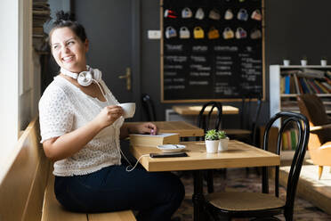 Lächelnde Frau, die eine Kaffeetasse hält und durch das Fenster schaut, während sie am Tisch in einem Café sitzt - GIOF08791