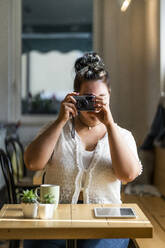 Junge Frau beim Fotografieren mit der Kamera, während sie am Tisch in einem Café sitzt - GIOF08798