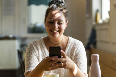 Nahaufnahme einer lächelnden, üppigen Frau, die ein Mobiltelefon benutzt, während sie im Restaurant sitzt - GIOF08801