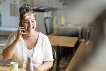 Fröhliche, üppige Frau, die in einem Café sitzt und über ein Mobiltelefon spricht - GIOF08805