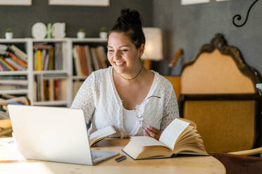 Smiling young woman studying over laptop by books on table in coffee shop - GIOF08806