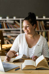 Smiling young woman writing in book while studying over laptop on table at coffee shop - GIOF08807