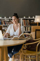 Lächelnde junge Frau mit Büchern und Laptop auf dem Tisch, die in einem Café studiert - GIOF08808