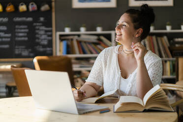 Voluptuous woman with books and laptop on table contemplating while sitting in cafe - GIOF08810