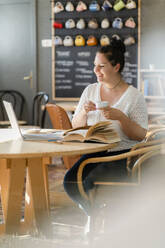 Smiling voluptuous woman holding coffee cup while studying over laptop in restaurant - GIOF08812