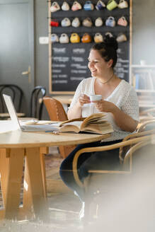 Lächelnde, üppige Frau, die eine Kaffeetasse hält, während sie über einem Laptop im Restaurant studiert - GIOF08812