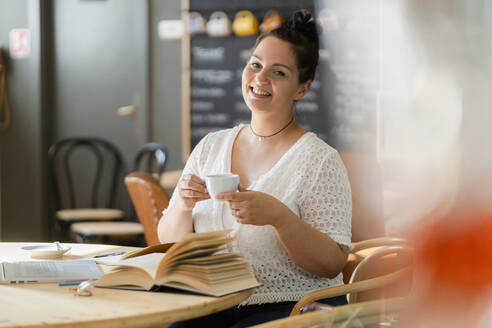 Lächelnde üppige Frau, die eine Kaffeetasse hält, während sie am Tisch im Restaurant sitzt - GIOF08813