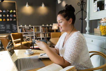 Young woman with books and laptop on table using smart phone in coffee shop - GIOF08814