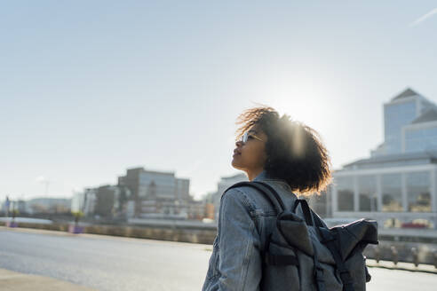 Young woman with backpack standing against clear sky in city during sunny day - BOYF01443
