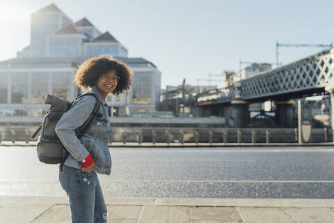 Smiling young woman with backpack standing on footpath by river in city stock photo