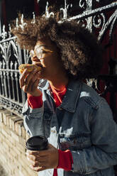 Nahaufnahme einer jungen Frau mit Afro-Haar, die an einem Zaun in der Stadt einen Donut isst - BOYF01445