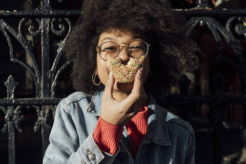 Close-up of young woman with curly hair eating donut against fence in city stock photo