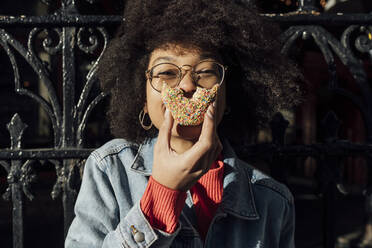 Close-up of young woman with curly hair eating donut against fence in city - BOYF01446