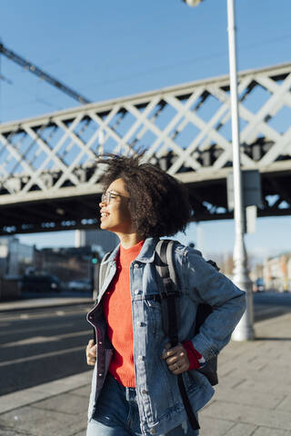 Smiling young woman with afro hair standing on sidewalk in city during sunny day stock photo