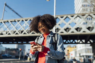 Lächelnde junge Frau mit Afro, die ein Smartphone benutzt, während sie an einer Brücke in der Stadt steht - BOYF01450