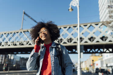 Fröhliche junge Frau mit Afrolook, die über ein Smartphone spricht, während sie an einer Brücke in der Stadt steht - BOYF01451