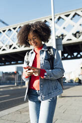 Afro junge Frau, die ein Smartphone benutzt, während sie auf dem Bürgersteig vor einer Brücke in der Stadt steht - BOYF01452