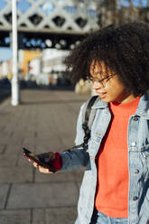 Nahaufnahme einer Frau mit lockigem Haar, die ein Mobiltelefon benutzt, während sie an einem sonnigen Tag auf dem Gehweg steht - BOYF01454