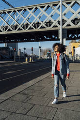 Junge Frau mit lockigem Haar, die auf dem Bürgersteig gegen eine Brücke in der Stadt läuft - BOYF01456