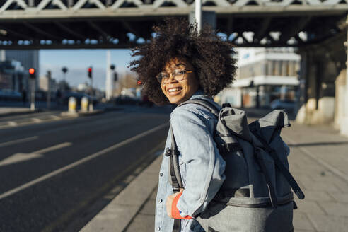 Lächelnde Afro-Frau mit Rucksack steht auf dem Bürgersteig in der Stadt an einem sonnigen Tag - BOYF01457