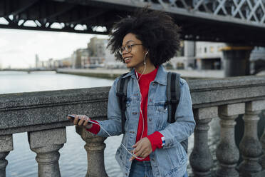 Junge Afroamerikanerin hört Musik über Kopfhörer, während sie am Geländer einer Fußgängerbrücke steht - BOYF01463