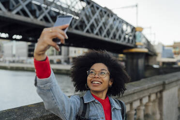 Lächelnde Afro-Frau, die ein Selfie mit ihrem Smartphone an einem Geländer in der Stadt macht - BOYF01465