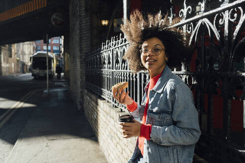 Junge Frau mit Afro-Haar hält Donut und Kaffee, während sie am Zaun in der Stadt steht - BOYF01472
