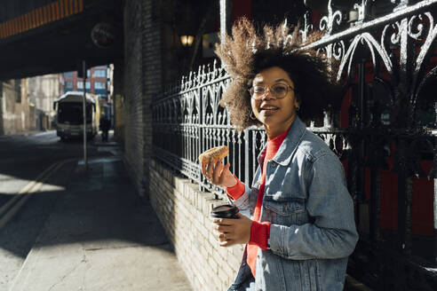 Young woman with afro hair holding donut and coffee while standing by fence in city - BOYF01472