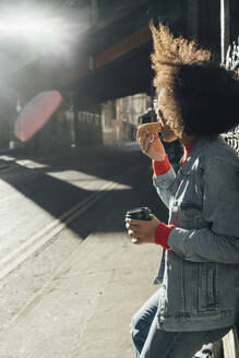 Junge Afro-Frau isst Donut, während sie an einem sonnigen Tag auf der Straße steht - BOYF01474