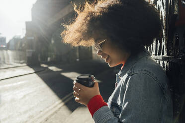 Nahaufnahme einer lächelnden Afro-Frau, die an einem sonnigen Tag einen Einwegbecher auf der Straße hält - BOYF01476