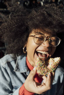 Close-up of afro young woman holding donut laughing outdoors during sunny day - BOYF01478