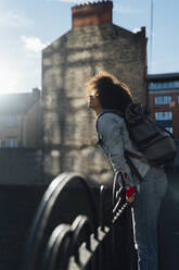 Unbekümmerte Afro-Frau mit Rucksack am Geländer stehend an einem sonnigen Tag - BOYF01484