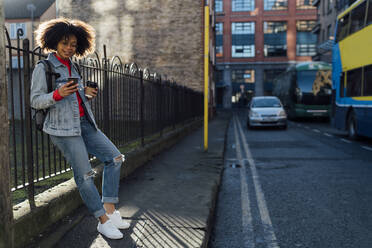 Afro young woman holding coffee cup using mobile phone while standing on street - BOYF01487