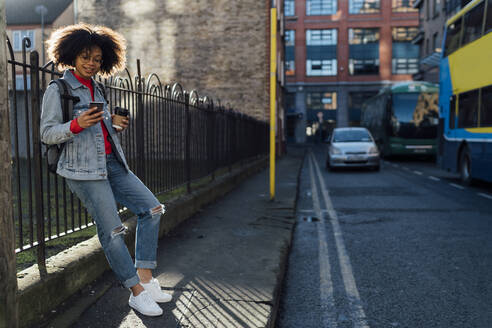 Junge Afro-Frau mit Kaffeetasse und Mobiltelefon auf der Straße stehend - BOYF01487